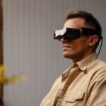 Person seated indoors wearing a Bigscreen Beyond 2 VR headset with its tethered cable
