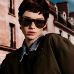 Model wearing olive-framed Ray-Ban Meta Gen 3 AI glasses with tinted lenses in front of a Paris building
